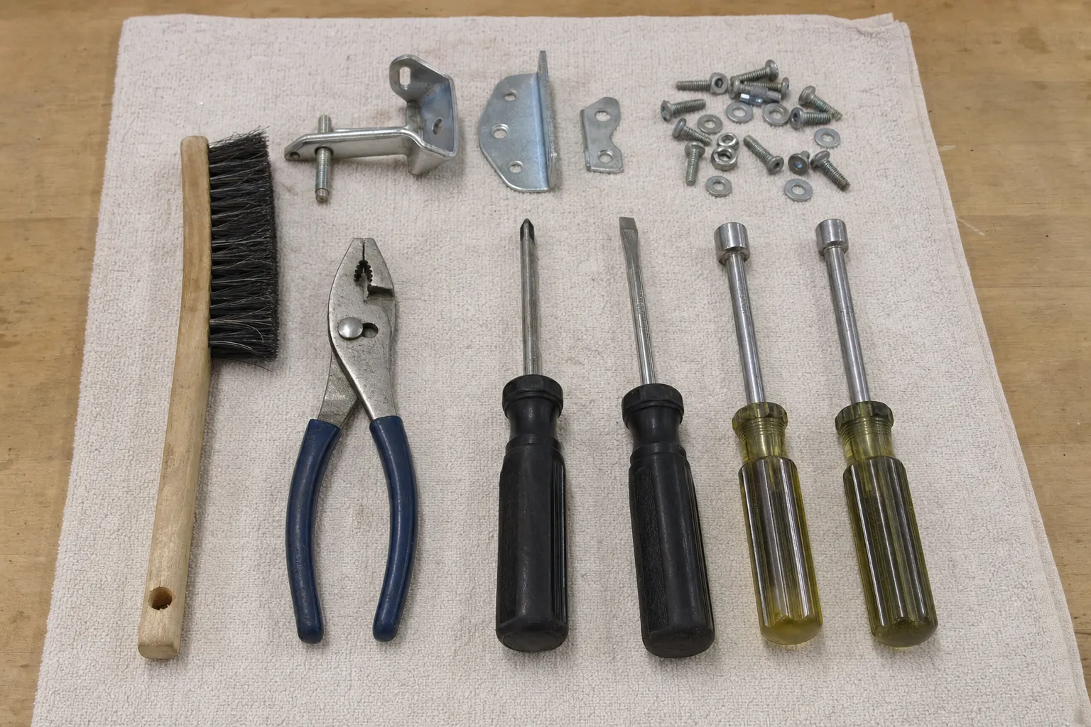 Wooden brush, blue-handled pliers, two screwdrivers and two nut drivers in a row on a cloth below three brackets, bolts, nuts and washers